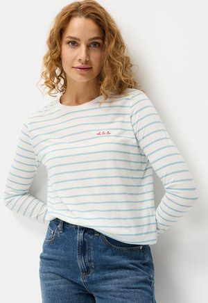 Langärmliges Hemd in Weiß mit hellblauen Streifen, mit einem kleinen roten gestickten Text "oh la la" auf der Brust. Kombiniert mit blauen Jeans.