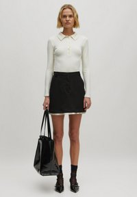 Weißes Langarmoberteil mit Kragen und Knopfdetails, kombiniert mit einem schwarzen Minirock mit kontrastierendem Saum. Schwarze Tragetasche und Absätze.