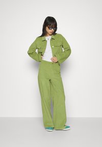 Groene fluwelen jas met bijpassende wijde broek, gestyled over een wit t-shirt. De outfit heeft knoopdetails en hoekige zonnebrillen.