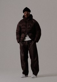 Homme portant un bonnet noir, des lunettes, une veste matelassée marron foncé, un pantalon marron foncé ample, des baskets blanches, se tenant devant un fond neutre.