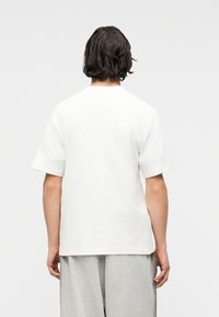 Άτομο με σκούρα μαλλιά, γυρισμένο προς τα πίσω, φοράει απλό λευκό κοντομάνικο μπλουζάκι και ανοιχτό γκρι χαλαρό παντελόνι μπροστά σε απλό φόντο.