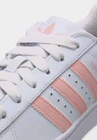 Λευκό αθλητικό παπούτσι με ροζ ρίγες, λευκά κορδόνια και ροζ λογότυπο trefoil στη γλώσσα, που εμφανίζει λεπτομερή ραφές και οπές.