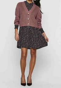 Rosa stickad cardigan med ribbad textur och tre knappar, bärs över en svart blommig klänning med utsvängd kjol och långa ärmar. Svarta klackar.