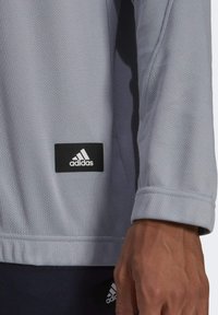 Lichtgrijs sportshirt van een gestructureerde stof, met lange mouwen, voorzien van een zwart rechthoekig adidas-logo label nabij de zoom.