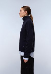 Marineblauer Rollkragenpullover mit lockerer Passform, aus weichem Material, mit gerippten Bündchen und einem dezenten Logo am Saum. Über einem helleren Hemd getragen.