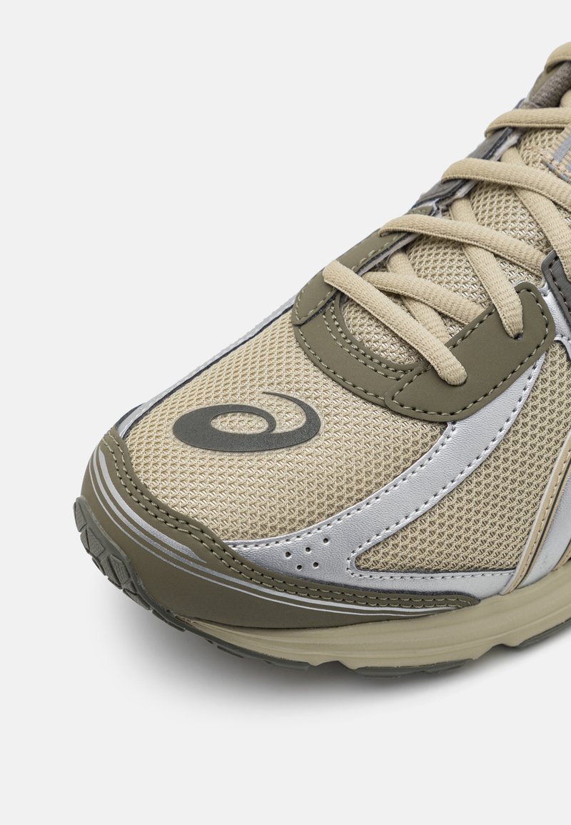 Zapato deportivo en tonos beige y oliva con parte superior de malla y acentos plateados, logotipo redondo y suela de goma texturizada.