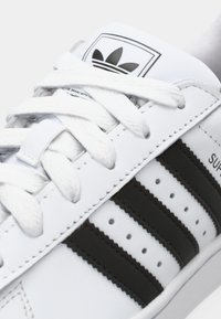 Close-up of white Adidas Superstar sneaker with black stripes, white laces, and Adidas trefoil logo on the tongue.