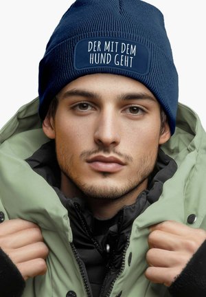 Navy knitted beanie with a white patch featuring text, worn by a person in a green puffer jacket with a black inner layer.