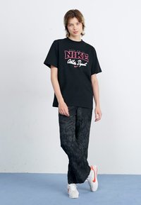 Black cotton t-shirt with red Nike logo, paired with black patterned pants, and white sneakers with red accents.