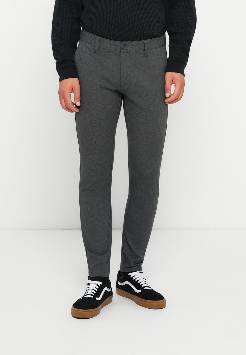 Pantalones ajustados grises hechos de una tela texturizada, con cierre de botón, trabillas para cinturón y bolsillos laterales, combinados con zapatillas negras.