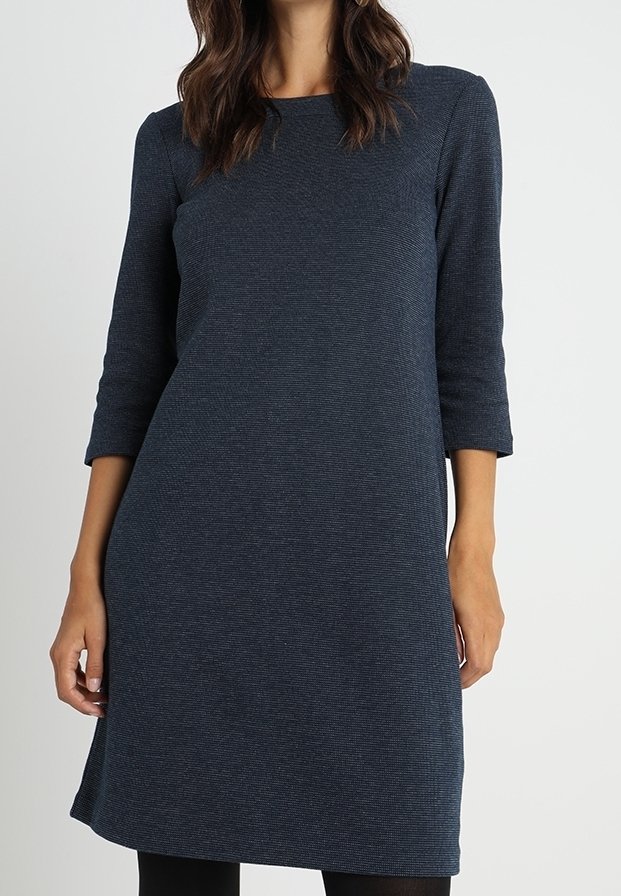 Femme portant une robe bleu foncé, longueur genou, manches longues, avec un col rond, sur un fond clair uni.