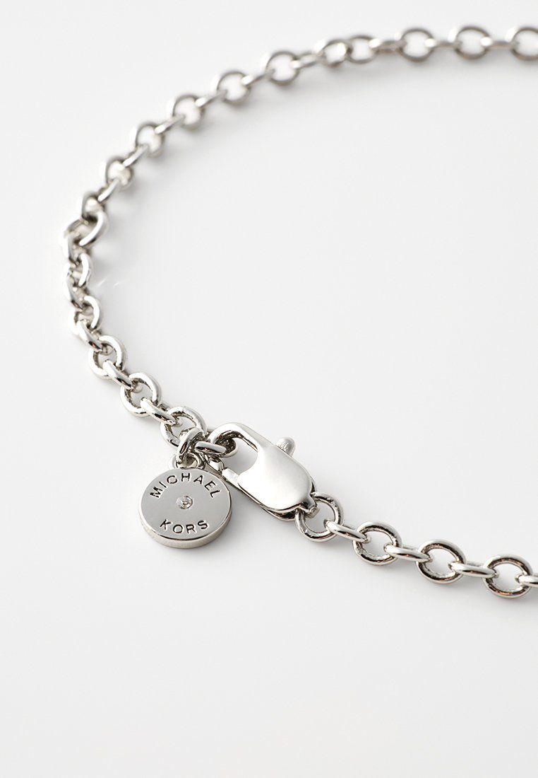 Srebrna bransoletka łańcuszkowa z okrągłą zawieszką z napisem "MICHAEL KORS", z zapięciem na zatrzask oraz okrągłymi ogniwami o gładkiej fakturze.