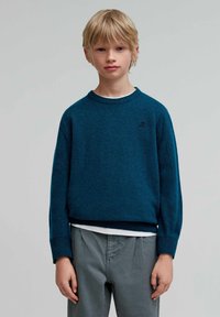 Suéter de punto en color verde azulado con cuello redondo y puños acanalados, combinado con pantalones grises. Presenta un logotipo sutil en el pecho.
