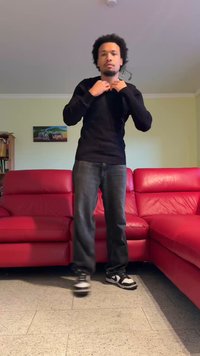 Jersey negro acanalado, vaqueros oscuros y zapatillas negras y blancas. De pie junto a un sofá de cuero rojo en una habitación con paredes de color claro.