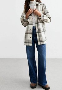 Camicia a quadri chiari con bottoni in tonalità crema e grigia, indossata sopra un dolcevita bianco, abbinata a jeans blu a vita alta e a gamba larga.