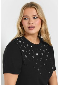 Camiseta negra de manga corta con adornos metálicos y de lentejuelas distribuidos en el frente, cuello redondo y una textura suave de algodón.