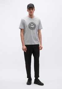 Pilkas trumpomis rankovėmis marškinėlis su juodu apskritu piešiniu, dėvimas kartu su juodomis kelnėmis ir juodais sportbačiais. Skrybėlė juoda su baltu logotipo užrašu.