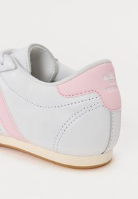 Baltas ādas sporta apavi ar rozā akcentiem, izšūtām detaļām un gumijas zoli. Aizmugurē ir logotips, kā arī gluda tekstūra.