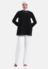 Top nero a maniche lunghe con scollatura rotonda, abbinato a pantaloni bianchi a gamba larga e tacchi a punta bianchi; include una collana di catena argentata.
