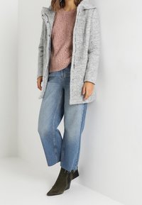 Cappotto in misto lana grigio con cappuccio, maglione lavorato a maglia rosa, jeans croppati azzurri chiari e stivali alla caviglia verde oliva. Tessuto testurizzato e vestibilità comoda.