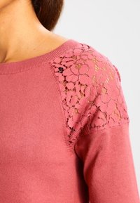 Růžový svetr s květinovými krajkovými detaily na ramenou, hladkou texturou a kulatým výstřihem. Detailní pohled na designové prvky.