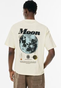 Kurze Ärmel, cremefarbenes T-Shirt mit einem blauen Mondmotiv, astronomischen Symbolen und schwarzem sowie braunem Text auf der Rückseite.