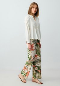 Blusa bianca a maniche lunghe con dettagli testurizzati, abbinata a pantaloni larghi con motivi floreali e foglie colorati. Sandali neutri.