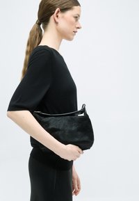 Γυναίκα με μακριά αλογοουρά που φοράει μαύρο τοπ και φούστα, κρατώντας μια μικρή μαύρη τσάντα με υφή, μπροστά σε φωτεινό φόντο.