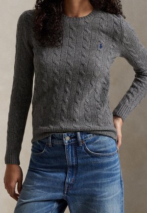 Femme portant un pull gris à torsades avec un petit logo bleu sur la poitrine et un jean bleu, debout avec une main sur la hanche.
