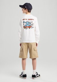 Long-sleeve white T-shirt featuring a colorful graphic of a skater on a skateboard with "KEITH HARING" text; paired with beige cargo shorts.