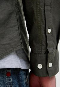 Camicia verde a maniche lunghe con bottoni bianchi, dettagli di cuciture, e orlo curvato, sovrapposta a una maglietta grigia.