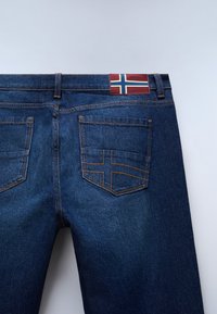 Jeansy dżinsowe w ciemnym niebieskim kolorze z pomarańczowym przeszyciem. Posiadają etykietę z poziomym i pionowym wzorem pasków w kolorach czerwonym, białym i niebieskim.