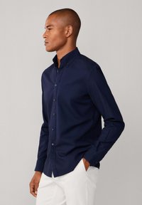 Camicia blu navy con bottoni in tessuto liscio, caratterizzata da un colletto strutturato e maniche lunghe, abbinata a pantaloni bianchi.