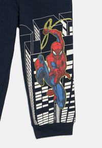 Spider-Man huśtający się przez miejską scenerię na ciemnoniebieskiej tkaninie, obrysowany na żółto, jedną ręką wystrzeliwujący sieć, a w tle budynki.