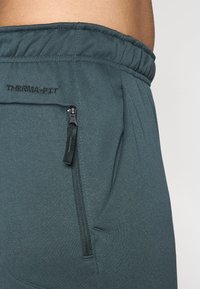 Zbliżenie na ciemnozielone dresy sportowe z elastycznym pasem, zamykaną kieszenią i etykietą "THERMA-FIT" z boku.