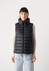 Gilet matelassé noir avec un col montant, fermeture éclair à l'avant et design matelassé horizontal. Porté par-dessus un haut à manches longues de couleur claire et un jean.