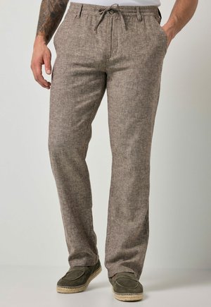 Hombre con pantalones de lino marrón claro con cintura ajustable, manos en los bolsillos y zapatos sin cordones verde oliva con suelas de cuerda.