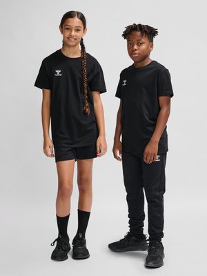 Zwei Kinder stehen, tragen schwarze Sportbekleidung mit Logos, Mädchen in Shorts und Junge in Hosen, beide in schwarzen Turnschuhen und Socken.