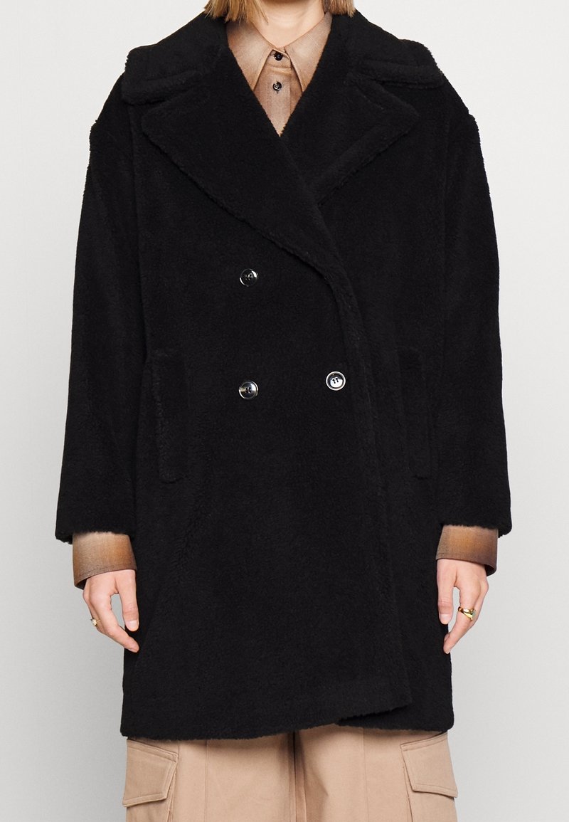 Manteau noir à double boutonnage en matière douce et texturée, avec de grands revers, des poches latérales et des boutons, porté par-dessus une chemise marron clair.