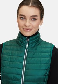 Grön vadderad väst med hög krage, framdragkedja och kontrasterande blå detaljer. Texturerad tyg med horisontella sömnadsdetaljer.