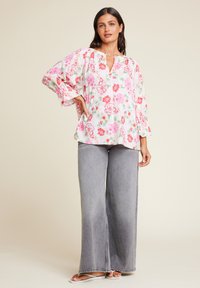 Hvit bluse med blomster i rosa og rødt, lange ermer med utsvingte mansjetter, kombinert med løse grå jeans og sandaler.