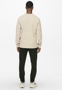 Sudadera de textura beige con mangas largas y cuello redondo, combinada con pantalones oscuros verdes de corte tapered y zapatillas blancas.