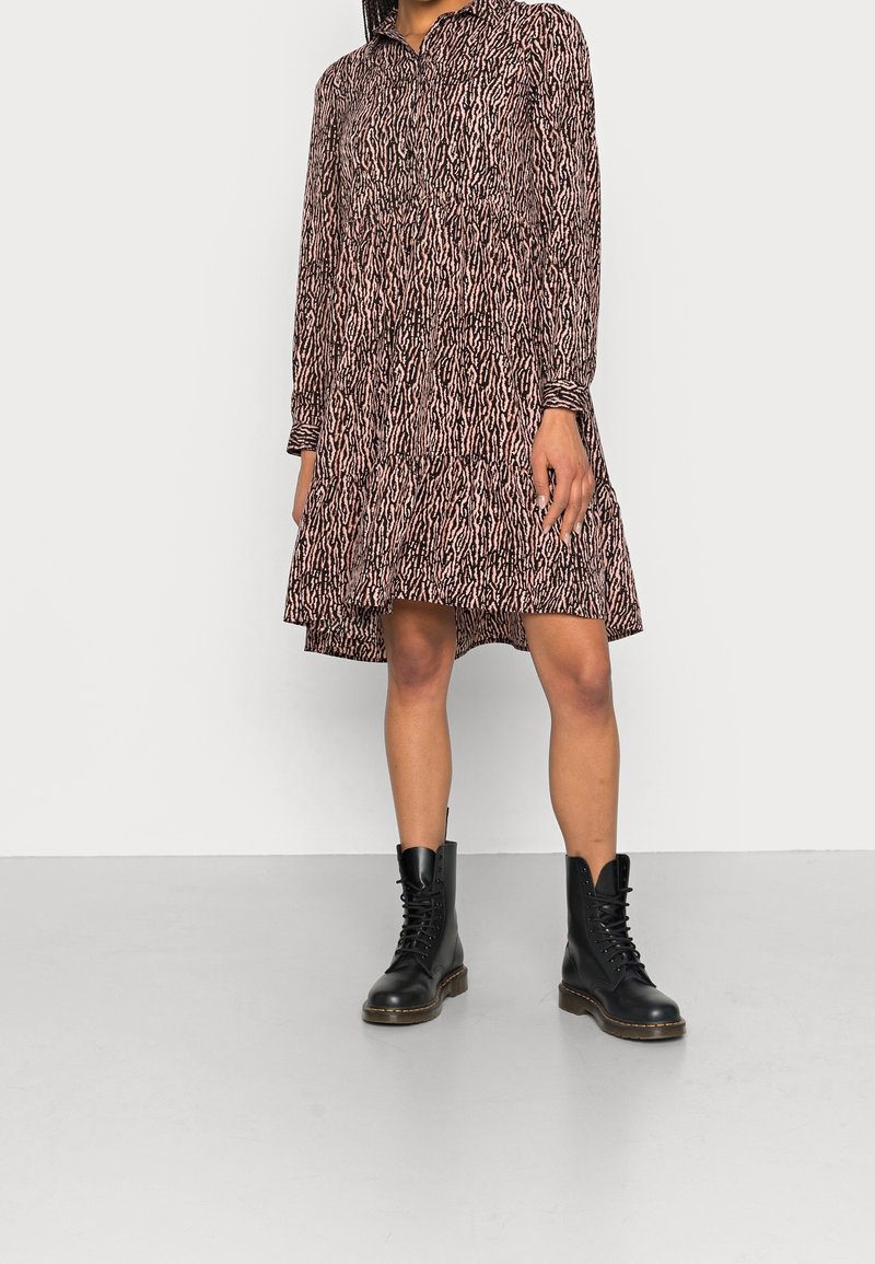 Robe à manches longues, longueur genou, avec un motif abstrait rose et noir, portée avec des bottes de combat noires à lacets sur un fond uni.