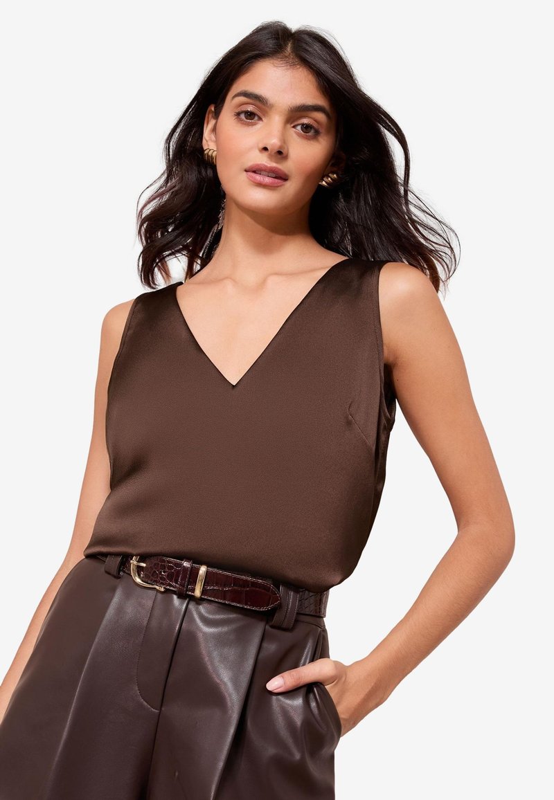 Donna con capelli scuri che indossa un top marrone senza maniche con scollo a V, pantaloni in pelle marrone scuro e una cintura abbinata con fibbia dorata, mano in tasca.
