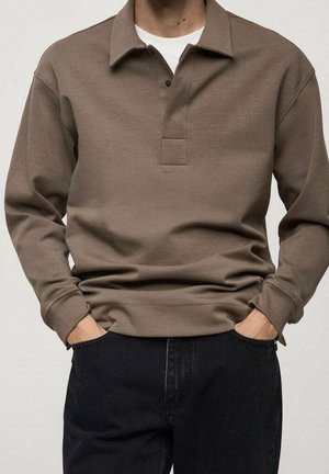 Man met een bruin poloshirt met lange mouwen over een wit onderhemd, handen in de zakken van een zwarte broek, staand tegen een effen achtergrond.