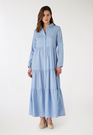 Donna in piedi che indossa un vestito lungo azzurro chiaro con bottoni, a balze, e scarpe beige con zeppa, su uno sfondo bianco semplice.