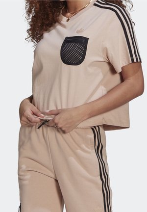 Femme portant une tenue beige Adidas avec trois bandes noires sur les manches et le pantalon, une poche noire zippée sur la chemise, ajustant la taille.