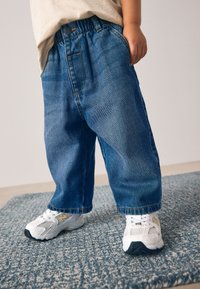 Pantaloni din denim cu talie elastică, croială lejeră și culoare albastră. Purtate cu adidași albi cu accente aurii. Suprafeță plată dedesubt.
