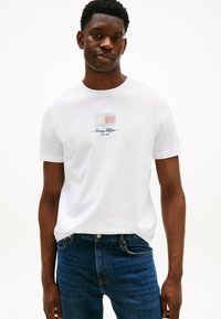 Hvid bomulds t-shirt med rund halsudskæring, der har et farverigt logo-design på brystet. Sammen med mørkeblå denim jeans.