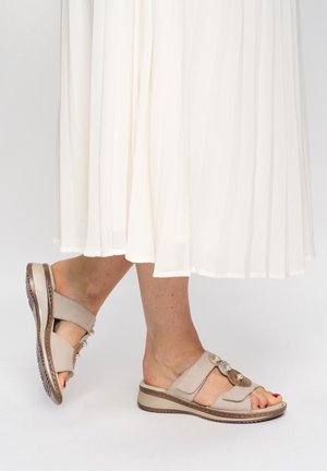 Feet wearing beige sandals with decorative pearls and embroidery, paired with a white pleated skirt on a light background.
